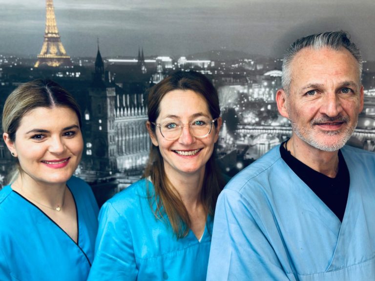 Drei Personen in blauen Kitteln, mit Paris-Hintergrund und lächelnd.